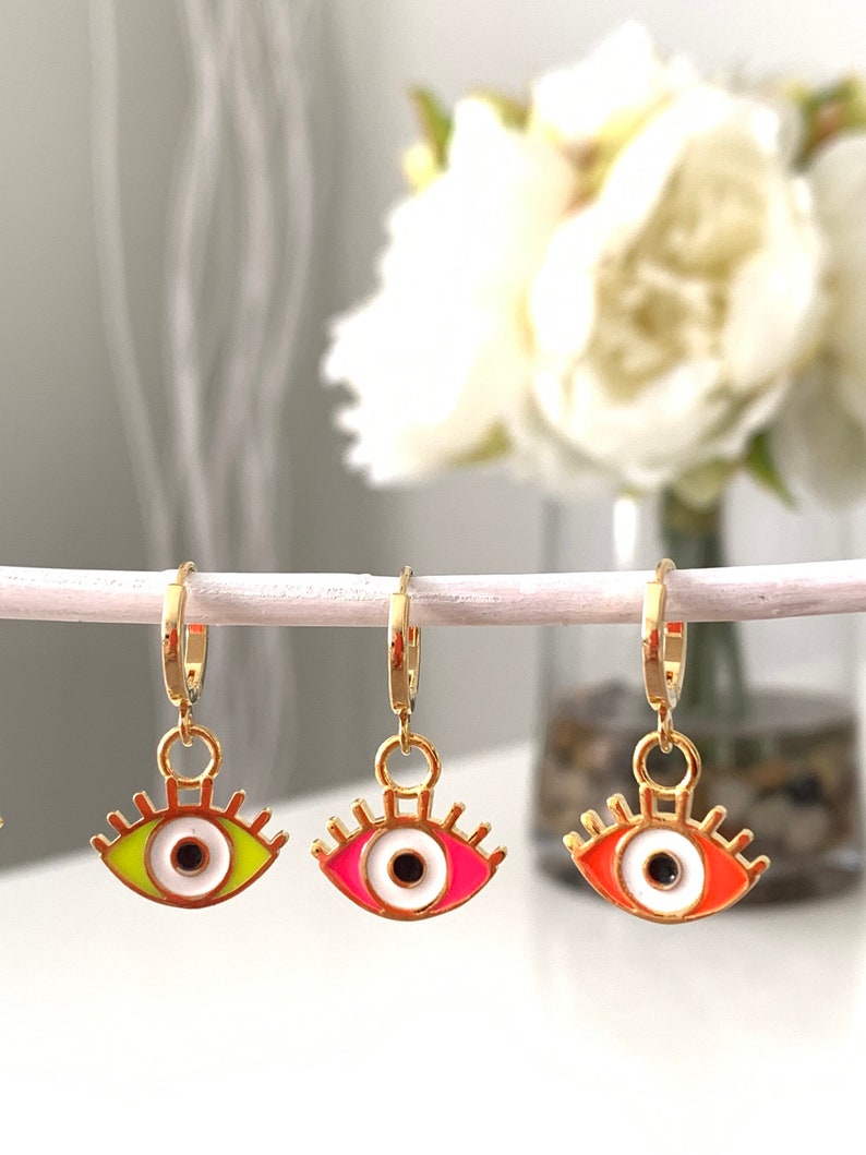 May include: Three gold hoop earrings with a dangling evil eye charm. The eyes are white with black pupils and have different colored rims: green, pink, and orange.