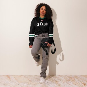 May include: A black long-sleeved jersey with green and white stripes on the sleeves. The jersey has the Arabic word "ليبان" printed in white on the front. A person is wearing the jersey with grey jeans and a black crossbody bag.