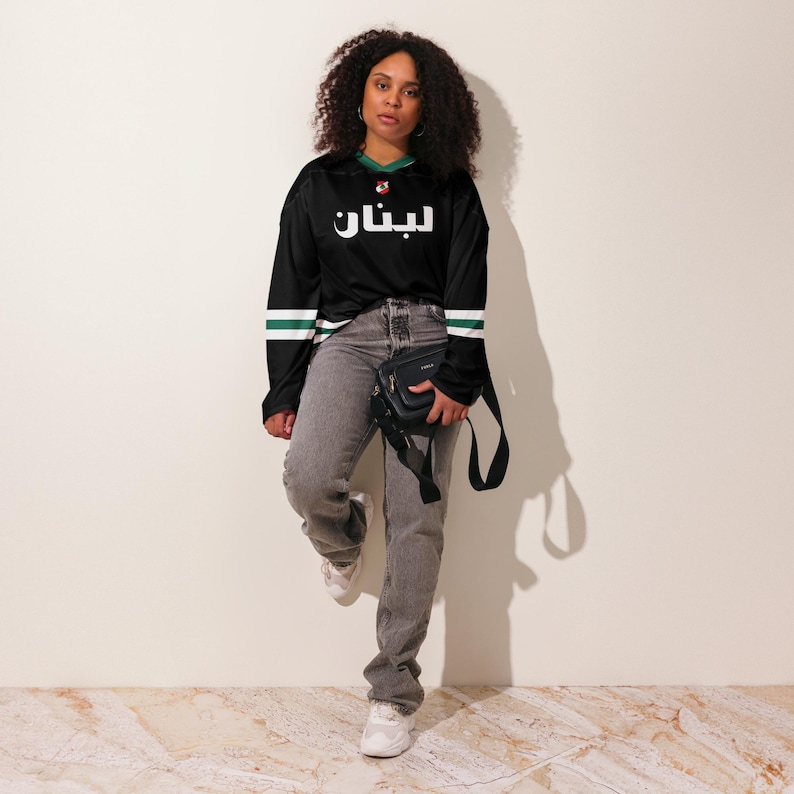 May include: A black long-sleeved jersey with green and white stripes on the sleeves. The jersey has the Arabic word "ليبان" printed in white on the front. A person is wearing the jersey with grey jeans and a black crossbody bag.