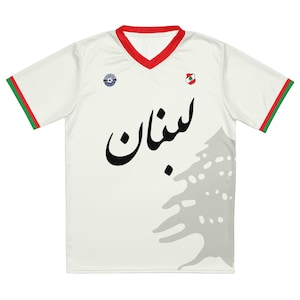 Op de afbeelding: Wit voetbaltenue met groene en rode afwerking op de mouwen. Het tenue heeft een grijze omtrek van een cederboom met het Arabische woord "Libanon" in het zwart op de voorkant. Het tenue heeft ook een klein logo op de linkerborst en een kleine Libanese vlag op de rechterborst.
