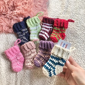 May include: A collection of handmade, miniature crochet Christmas stockings in various colors, including pink, green, blue, and red. Some stockings have decorative patterns. One stocking holds a gift card. The stockings are displayed on a soft, textured surface.
