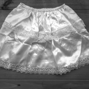 May include: A white satin skirt with lace trim. The skirt has a gathered waist and two tiers of fabric, with delicate lace edging the hem and tiers. The fabric has a subtle sheen, and the skirt is laid flat on a wooden surface.