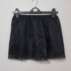 Puede incluir: Falda interior negra colgada de una percha negra. La falda está hecha de una tela brillante y tiene un delicado ribete de encaje a lo largo del dobladillo. La falda es de color negro liso.