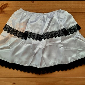 May include: A white satin skirt with three tiers of black lace trim. The skirt has an elastic waistband and a ruffled hem. The fabric has a slight sheen, and the lace adds a delicate contrast to the smooth satin.