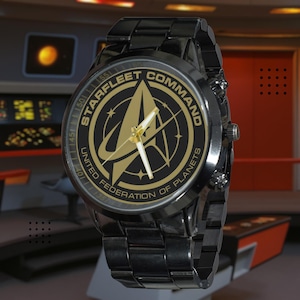 Peut inclure: Une montre-bracelet en métal noir avec un cadran rond arborant le logo doré Starfleet Command United Federation of Planets. Le cadran comprend des index dorés, des aiguilles dorées et un bracelet à maillons noirs. Le texte Starfleet Command et United Federation of Planets est imprimé en doré autour de l'emblème central.