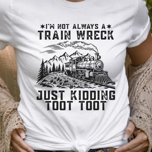 May include: White t-shirt with a black and white graphic of a train, mountains, and trees. Text on the shirt reads "I'm not always a train wreck, just kidding toot toot."