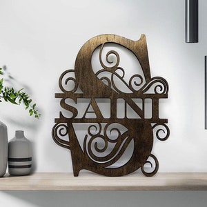 May include: Wooden monogram sign in a dark brown finish. The letter "S" is featured with the name "SAINI" cut out below. The design includes decorative swirls and flourishes, suitable for home decor.