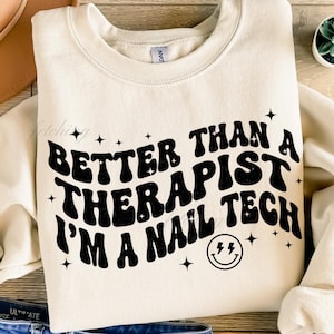 May include: Cream-colored sweatshirt with the text "BETTER THAN A THERAPIST I'M A NAIL TECH" in black, along with star and lightning bolt graphics. The sweatshirt has a crew neck and long sleeves.