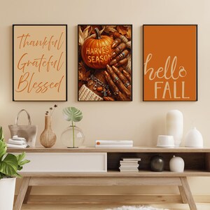 May include: A set of three framed wall art prints. The left print features the text Thankful Grateful Blessed in orange. The center print shows a pumpkin with the text HARVEST SEASON and hands with rings. The right print displays the text hello FALL.