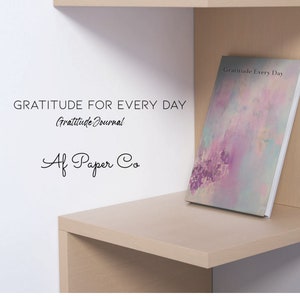 May include: A journal titled "Gratitude Every Day" with a soft, abstract cover design in shades of purple, pink, and blue. The journal is displayed on a light-colored wooden shelf, with text on the wall that reads "Gratitude for Every Day" and "Af Paper Co."