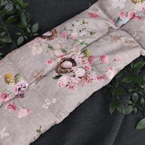 Könnte beinhalten: Ein graues und weißes Kissen mit einem Blumenmuster mit rosa Blumen, Schmetterlingen und Vogelnestern mit Eiern. Das Kissen ist aus einem weichen, gesteppten Stoff.