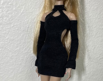 Può includere: Una bambola di moda che indossa un miniabito nero con tagli e maniche lunghe. La bambola ha lunghi capelli biondi e indossa sandali neri con tacco.