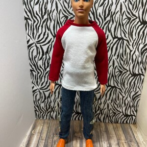 May include: A doll wearing a white and red baseball shirt, blue jeans with silver accents, and orange shoes. The doll has blonde hair and blue eyes.