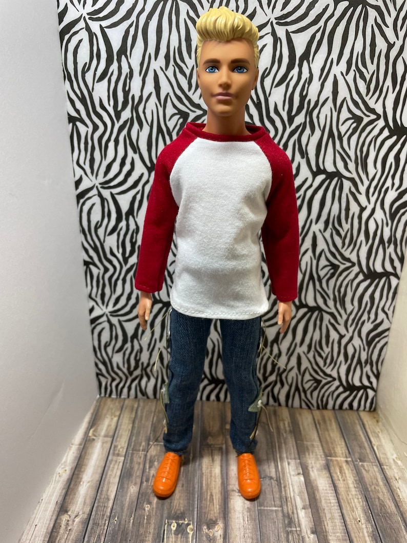 May include: A doll wearing a white and red baseball shirt, blue jeans with silver accents, and orange shoes. The doll has blonde hair and blue eyes.