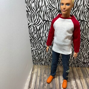 May include: A doll wearing a white and red baseball shirt, blue jeans, and orange shoes. The doll is standing in front of a zebra print background.