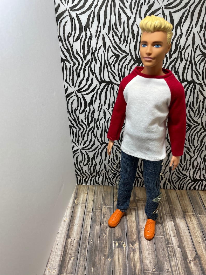 May include: A doll wearing a white and red baseball shirt, blue jeans, and orange shoes. The doll is standing in front of a zebra print background.
