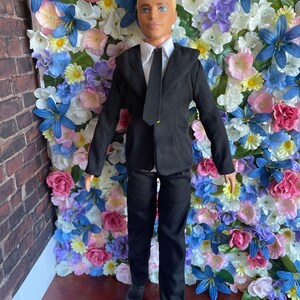 Puede incluir: Una mu&ntilde;eca que lleva un traje negro con una camisa blanca y una corbata negra. La mu&ntilde;eca tiene el pelo rubio y los ojos azules. La mu&ntilde;eca est&aacute; de pie frente a una pared de flores artificiales.