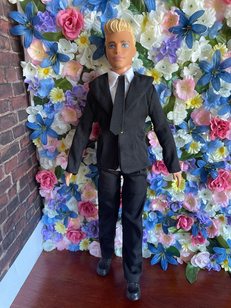 Puede incluir: Una mu&ntilde;eca que lleva un traje negro con una camisa blanca y una corbata negra. La mu&ntilde;eca tiene el pelo rubio y los ojos azules. La mu&ntilde;eca est&aacute; de pie frente a una pared de flores artificiales.