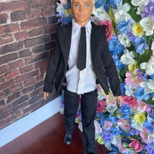 Puede incluir: Una mu&ntilde;eca que lleva un traje negro con una camisa blanca y una corbata negra. La mu&ntilde;eca tiene el pelo rubio y los ojos azules. La mu&ntilde;eca est&aacute; de pie frente a una pared de flores artificiales.