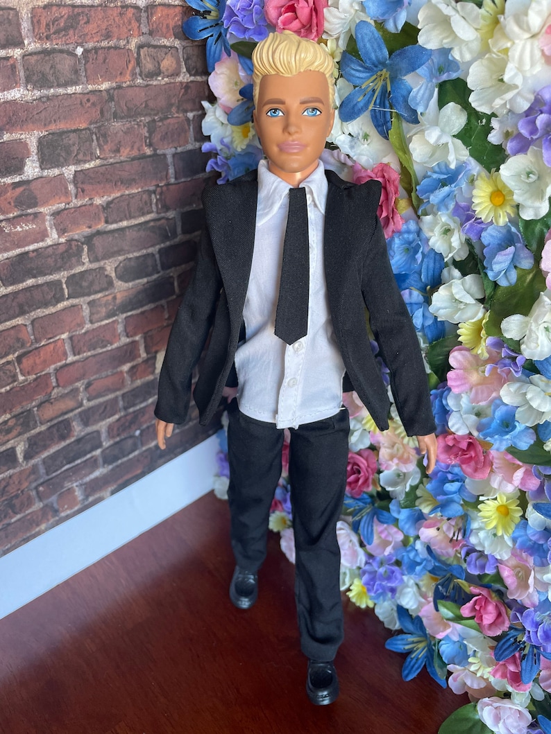 Puede incluir: Una mu&ntilde;eca que lleva un traje negro con una camisa blanca y una corbata negra. La mu&ntilde;eca tiene el pelo rubio y los ojos azules. La mu&ntilde;eca est&aacute; de pie frente a una pared de flores artificiales.