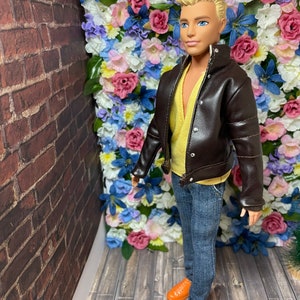 May include: A doll wearing a brown leather jacket, a yellow shirt, blue jeans, and orange shoes. The doll is standing in front of a wall of artificial flowers.