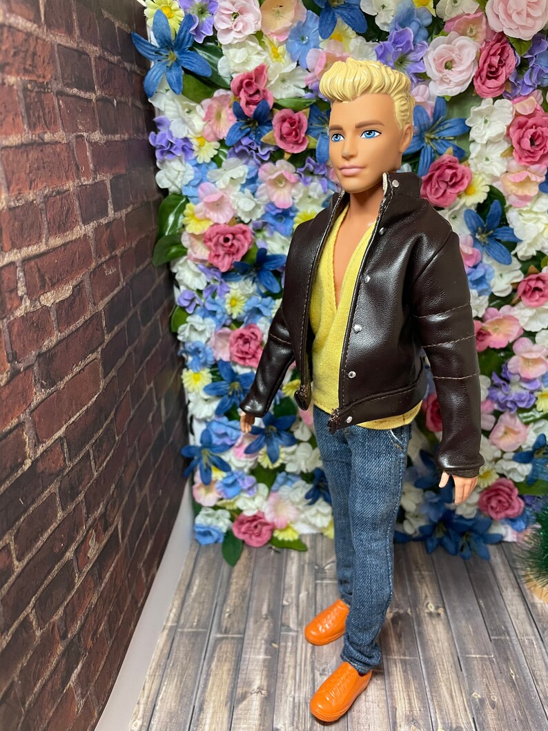 May include: A doll wearing a brown leather jacket, a yellow shirt, blue jeans, and orange shoes. The doll is standing in front of a wall of artificial flowers.