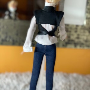 May include: A doll wearing a white shirt with a black vest, blue jeans, and blonde hair.