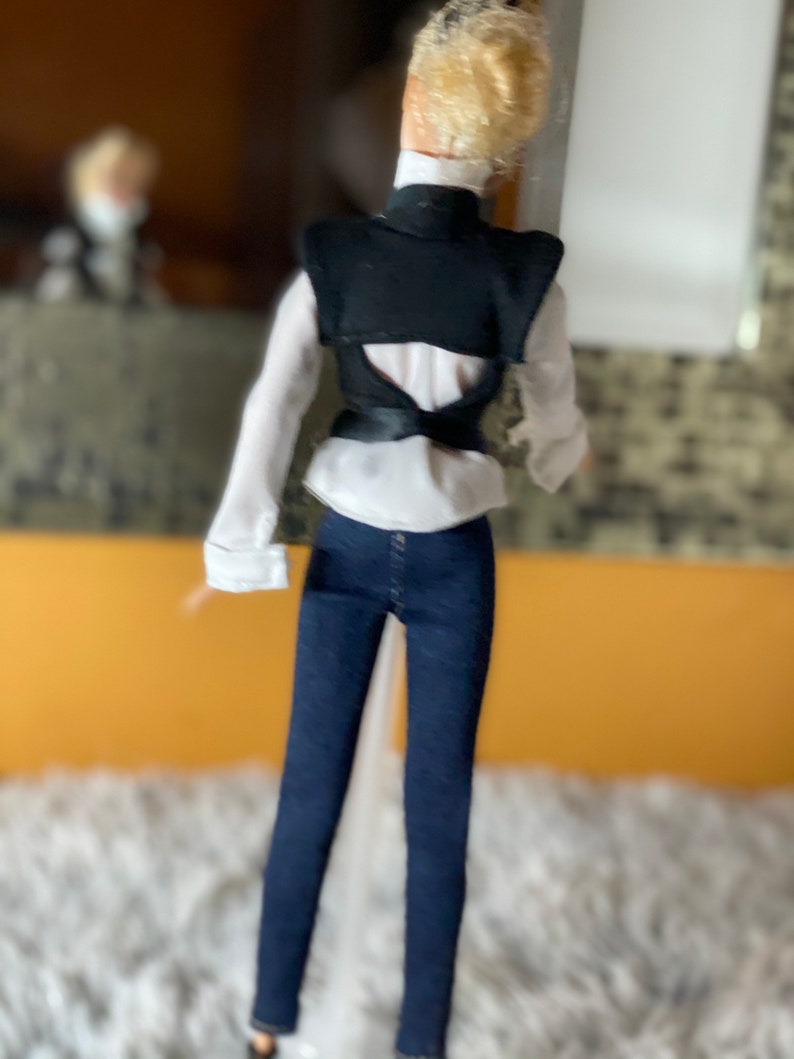 May include: A doll wearing a white shirt with a black vest, blue jeans, and blonde hair.