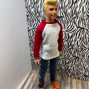 May include: A doll wearing a white and red baseball shirt, blue jeans, and orange shoes. The doll has blonde hair and blue eyes.