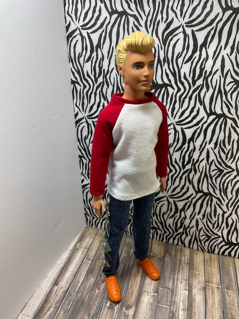 May include: A doll wearing a white and red baseball shirt, blue jeans, and orange shoes. The doll has blonde hair and blue eyes.