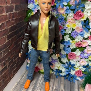 May include: A doll wearing a brown leather jacket, a yellow shirt, blue jeans, and orange shoes. The doll is standing in front of a wall of artificial flowers.