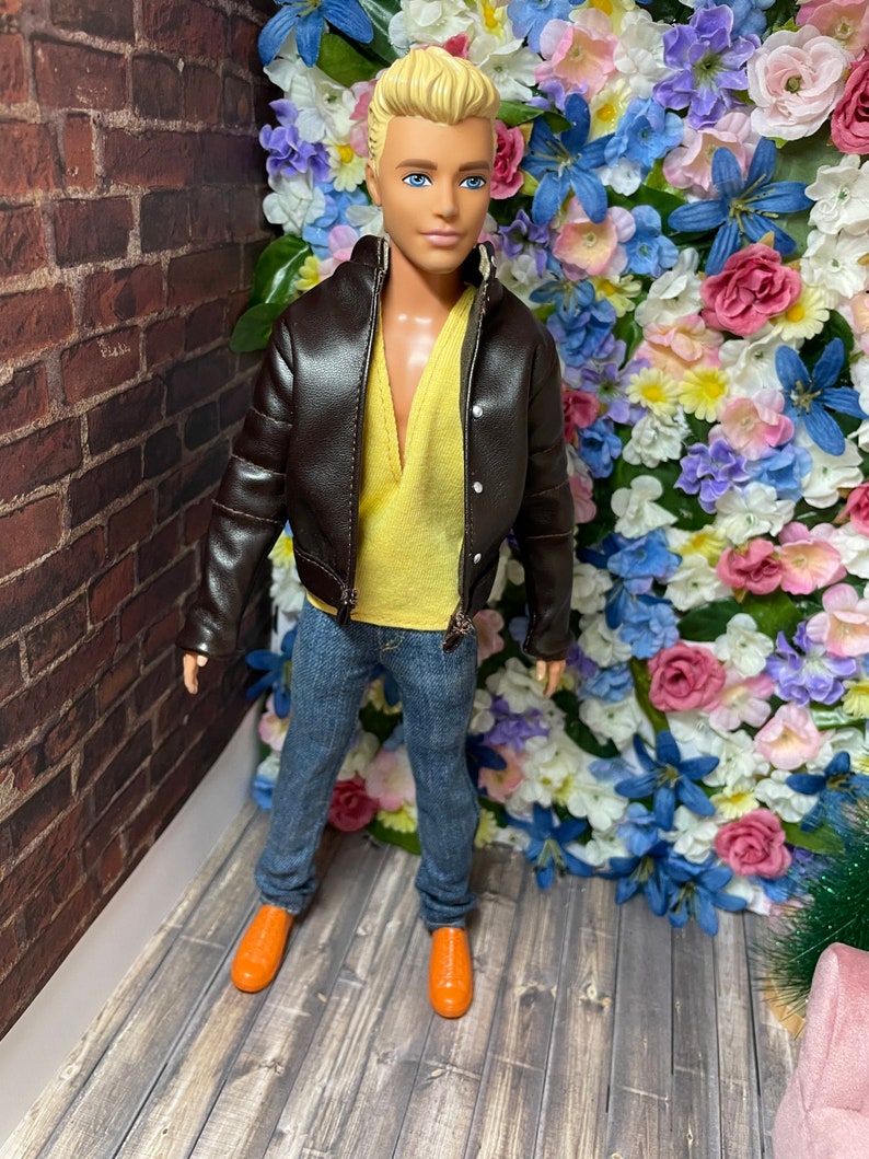 May include: A doll wearing a brown leather jacket, a yellow shirt, blue jeans, and orange shoes. The doll is standing in front of a wall of artificial flowers.