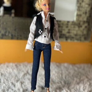 May include: A blonde doll wearing a white button-down shirt with a black vest, blue jeans, and black heels.