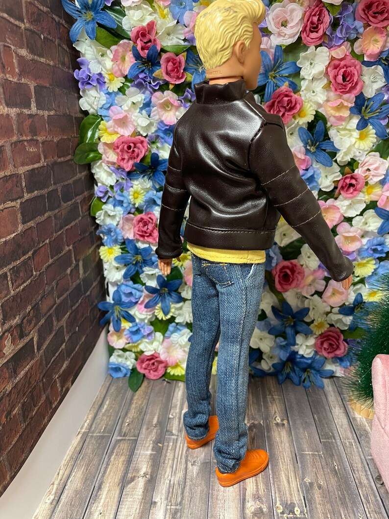 May include: A doll wearing a brown leather jacket, blue jeans, and orange shoes. The doll is standing in front of a wall of artificial flowers.