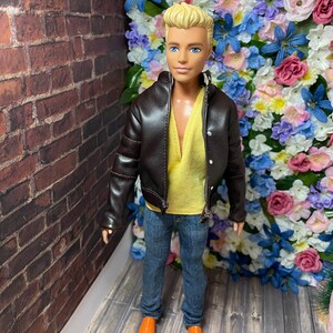 May include: A doll wearing a brown leather jacket, a yellow shirt, blue jeans, and orange shoes. The doll is standing in front of a wall of artificial flowers.