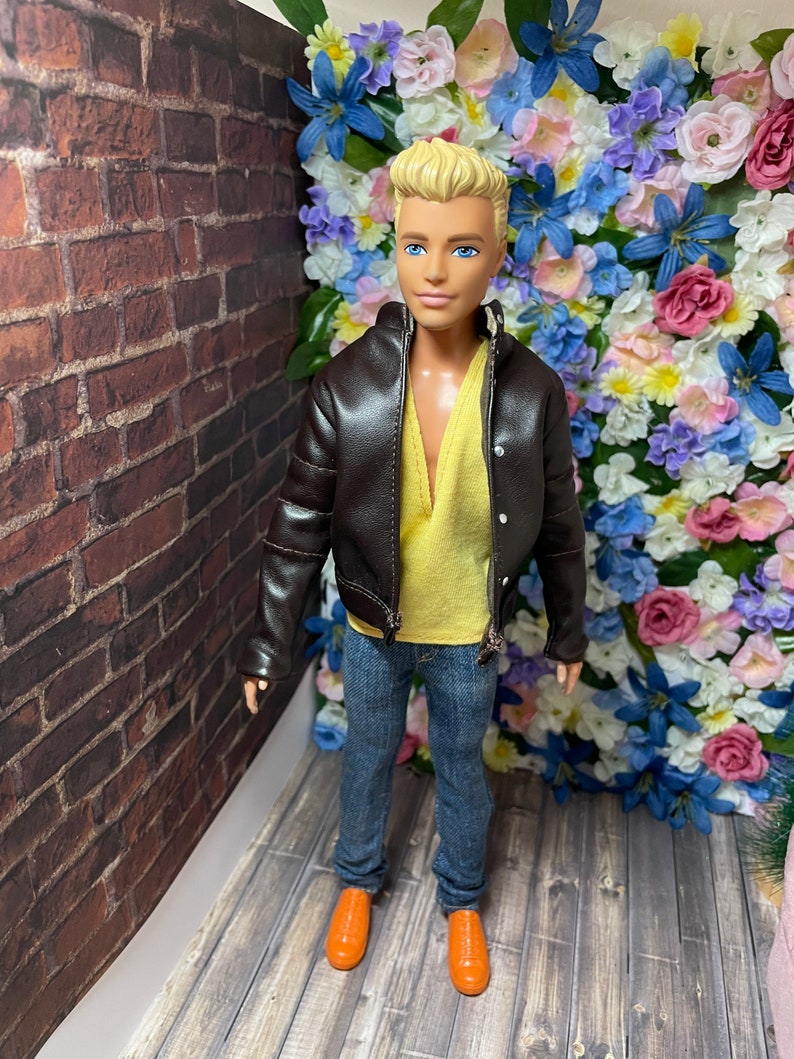 May include: A doll wearing a brown leather jacket, a yellow shirt, blue jeans, and orange shoes. The doll is standing in front of a wall of artificial flowers.