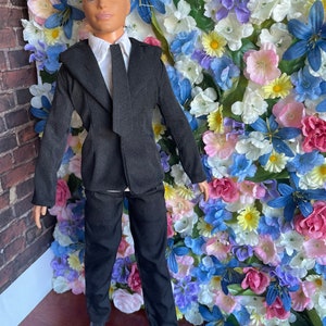 Puede incluir: Una mu&ntilde;eca vestida con un traje negro con camisa blanca y corbata negra. La mu&ntilde;eca tiene el pelo rubio y los ojos azules. La mu&ntilde;eca est&aacute; de pie frente a una pared de flores artificiales.