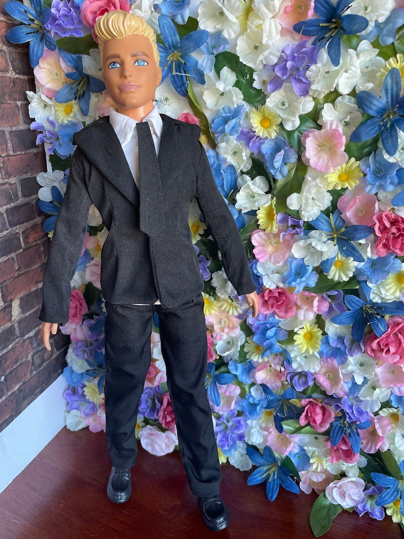Puede incluir: Una mu&ntilde;eca vestida con un traje negro con camisa blanca y corbata negra. La mu&ntilde;eca tiene el pelo rubio y los ojos azules. La mu&ntilde;eca est&aacute; de pie frente a una pared de flores artificiales.