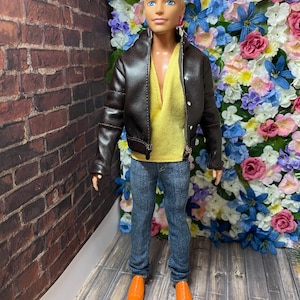 May include: A doll wearing a brown leather jacket, a yellow shirt, blue jeans, and orange shoes. The doll is standing in front of a wall of artificial flowers.