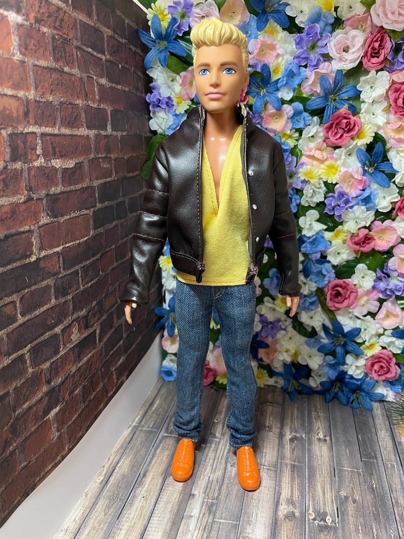 May include: A doll wearing a brown leather jacket, a yellow shirt, blue jeans, and orange shoes. The doll is standing in front of a wall of artificial flowers.