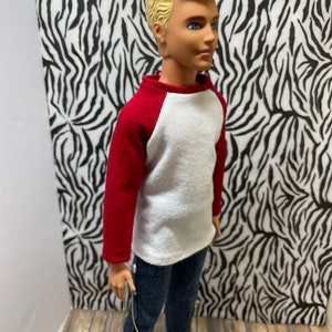 May include: A doll wearing a white and red baseball shirt, blue jeans, and orange shoes. The doll has blonde hair and blue eyes.