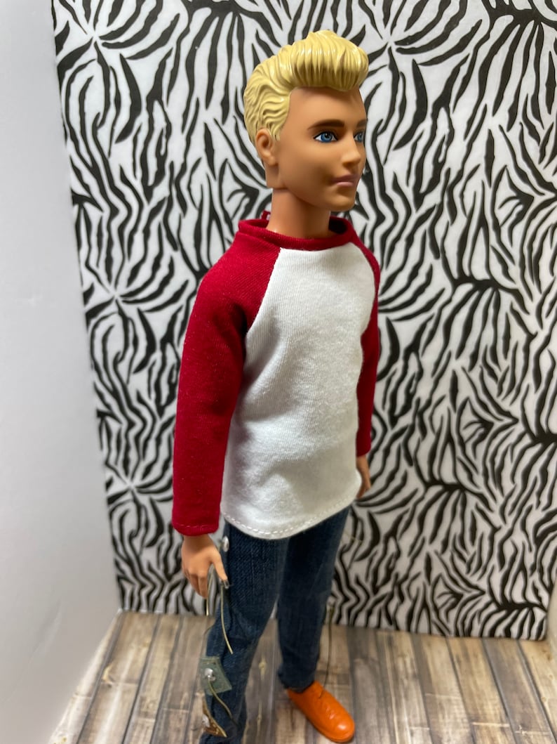 May include: A doll wearing a white and red baseball shirt, blue jeans, and orange shoes. The doll has blonde hair and blue eyes.