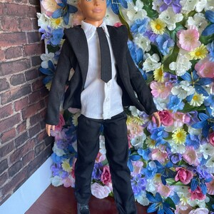 Puede incluir: Una mu&ntilde;eca vestida con un traje negro con camisa blanca y corbata negra. La mu&ntilde;eca tiene el pelo rubio y los ojos azules. La mu&ntilde;eca est&aacute; de pie frente a una pared de flores artificiales.