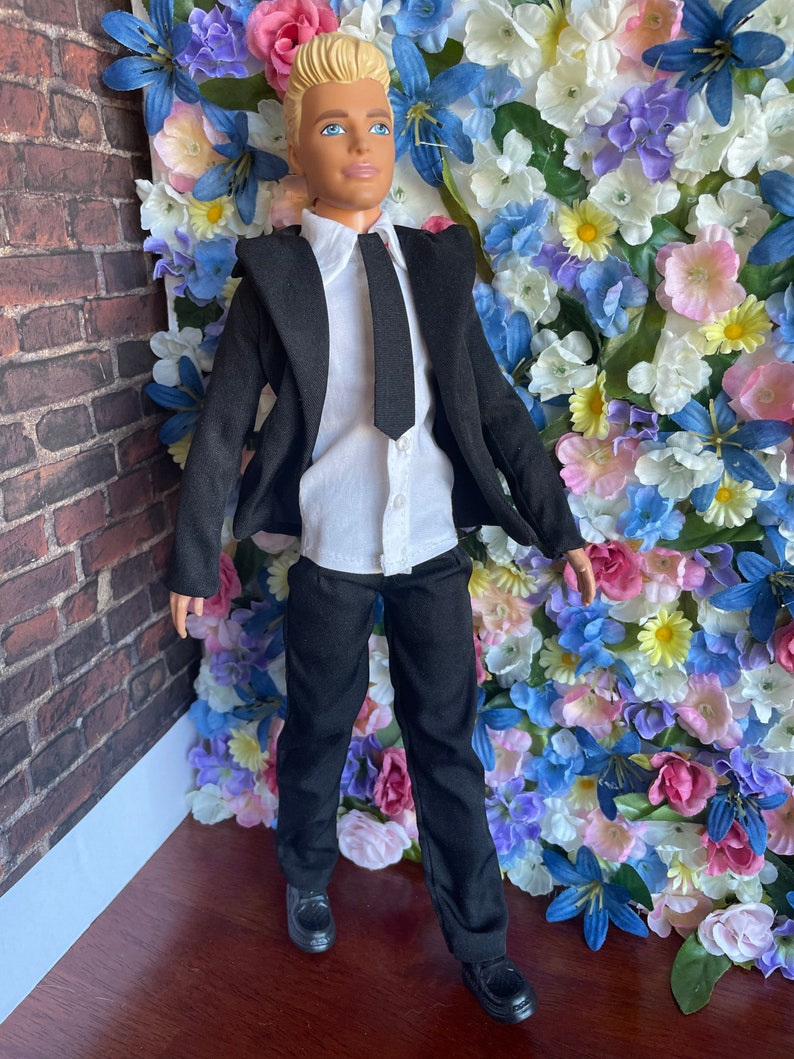 Puede incluir: Una mu&ntilde;eca vestida con un traje negro con camisa blanca y corbata negra. La mu&ntilde;eca tiene el pelo rubio y los ojos azules. La mu&ntilde;eca est&aacute; de pie frente a una pared de flores artificiales.