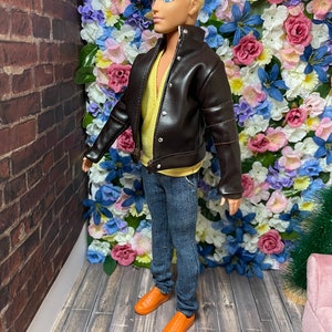 May include: A doll wearing a brown leather jacket, blue jeans, and orange shoes. The doll is standing in front of a wall of artificial flowers.