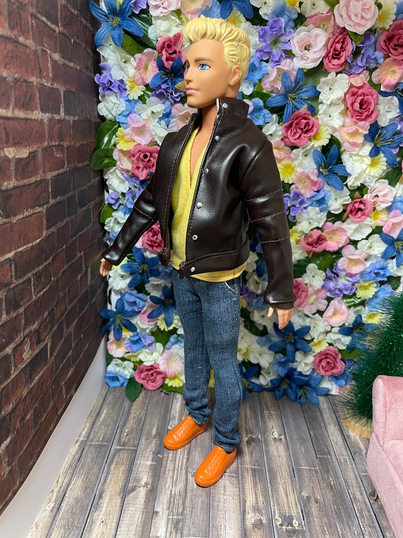 May include: A doll wearing a brown leather jacket, blue jeans, and orange shoes. The doll is standing in front of a wall of artificial flowers.