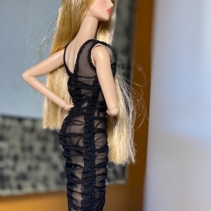 Pu&ograve; includere: Una bambola alla moda con lunghi capelli biondi e un abito a rete nero. L'abito &egrave; aderente e arricciato, con un design senza maniche. La bambola &egrave; in posa con un braccio di lato e l'altra mano sul fianco.