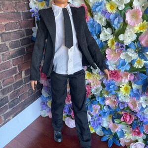 Puede incluir: Una mu&ntilde;eca vestida con un traje negro, camisa blanca y corbata negra. La mu&ntilde;eca est&aacute; de pie frente a una pared de flores artificiales.