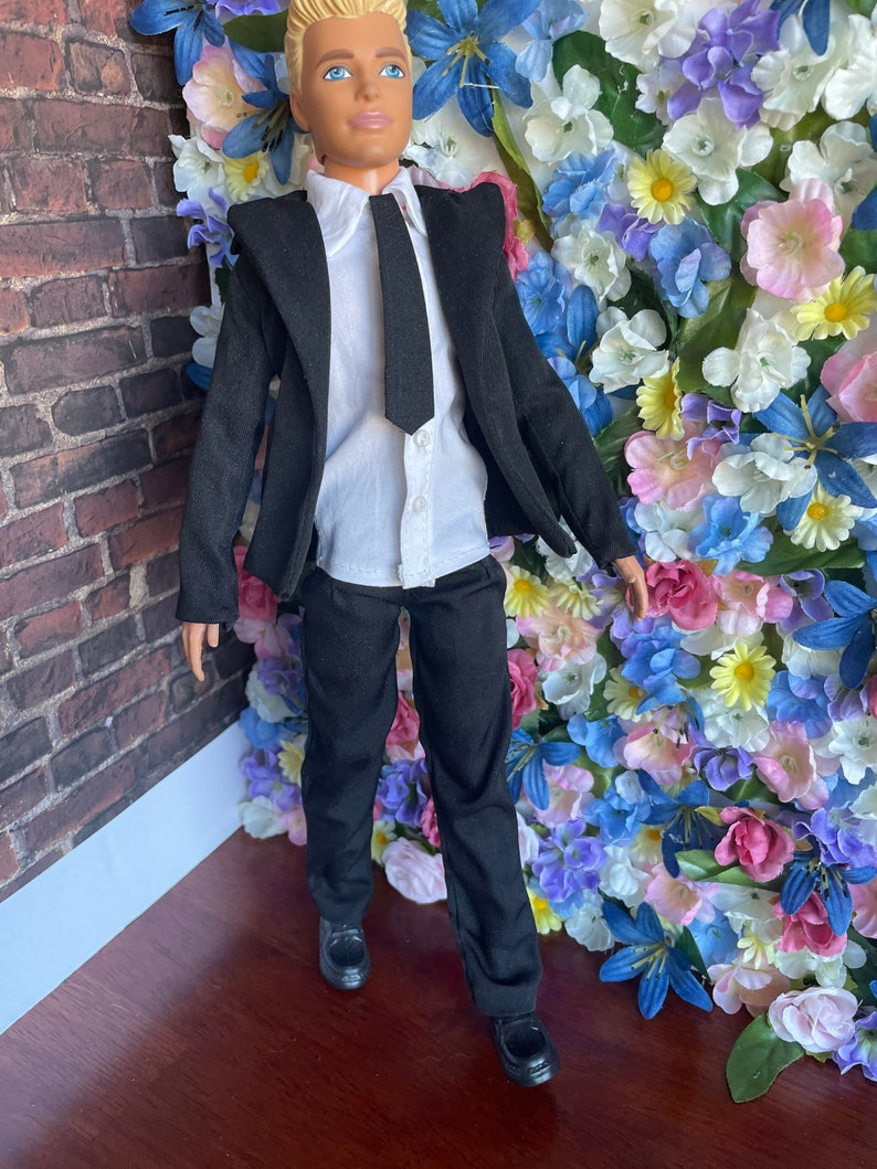 Puede incluir: Una mu&ntilde;eca vestida con un traje negro, camisa blanca y corbata negra. La mu&ntilde;eca est&aacute; de pie frente a una pared de flores artificiales.