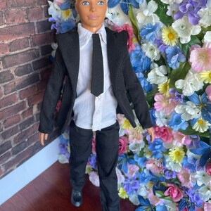 Puede incluir: Una mu&ntilde;eca que lleva un traje negro con una camisa blanca y una corbata negra. La mu&ntilde;eca tiene el pelo rubio y los ojos azules. La mu&ntilde;eca est&aacute; de pie frente a una pared de flores artificiales.
